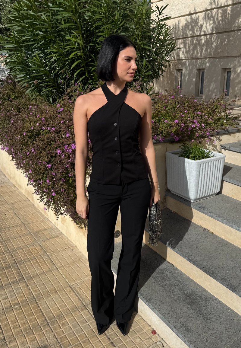 Femme avec une coupe au carré noire portant un haut halter noir, un pantalon noir et des chaussures pointues, tenant un sac à main perlé près de marches et de plantes.