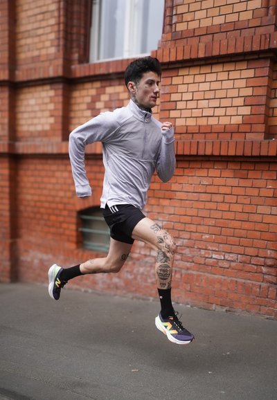 Camiseta de manga larga y cremallera de color gris claro, pantalones cortos negros y zapatillas de correr multicolores. Tatuajes visibles en las piernas. Corriendo por un camino de ladrillos al aire libre.