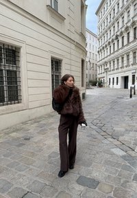 Rjava jakna iz umetnega krzna z dolgimi rokavi, v kombinaciji z temno rjavimi širokimi hlačami in črnimi čevlji. V roki drži pleteno črno torbico in skodelico kave.