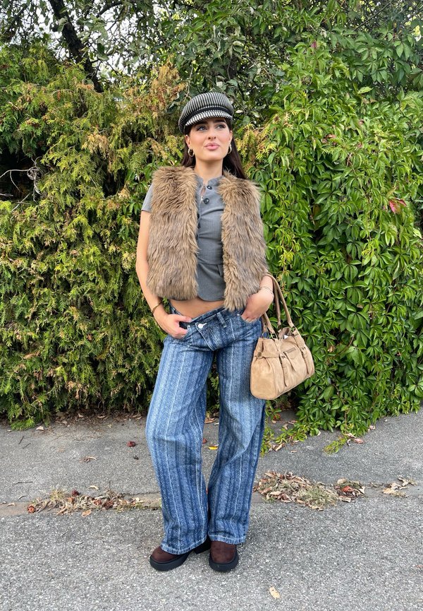Mujer con gorra de cuadros, chaleco de piel marrón, top corto gris, vaqueros azules a rayas y zapatos marrones, sosteniendo un bolso beige junto a arbustos verdes.