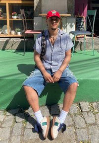 Indivíduo a usar uma camisa xadrez de mangas curtas, calções de ganga destruídos e sapatos multicoloridos, sentado numa plataforma verde com paralelepípedos.