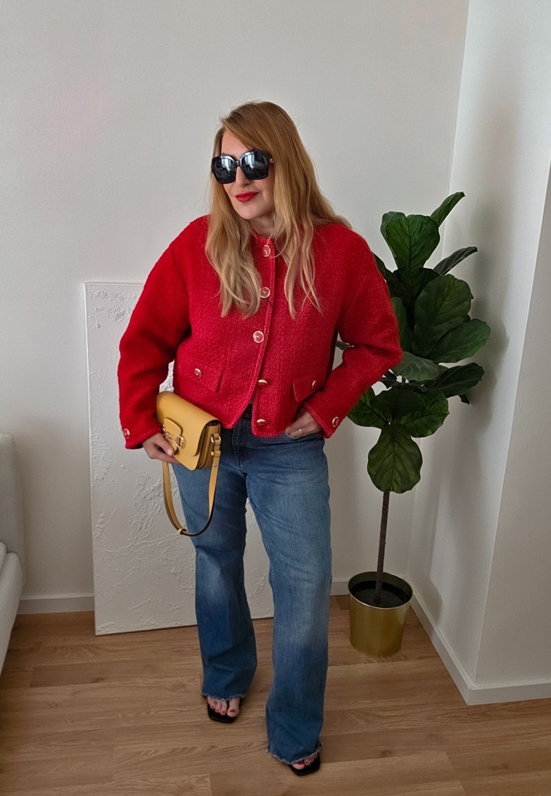 Kvinne iført en rød jakke og blå jeans holder en sennepsfarget håndveske, stående ved siden av en grønn potteplante i et rom med tregulv.