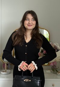 Jeune femme aux longs cheveux noirs portant une robe noire boutonnée et un collier de perles, tenant un sac à main noir, debout dans une pièce avec des fleurs.
