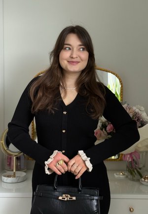Joven con cabello largo y oscuro, vistiendo un vestido negro abotonado y un collar de perlas, sosteniendo un bolso negro, de pie en una habitación con flores.