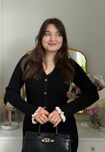 Joven con cabello largo y oscuro, vistiendo un vestido negro abotonado y un collar de perlas, sosteniendo un bolso negro, de pie en una habitación con flores.