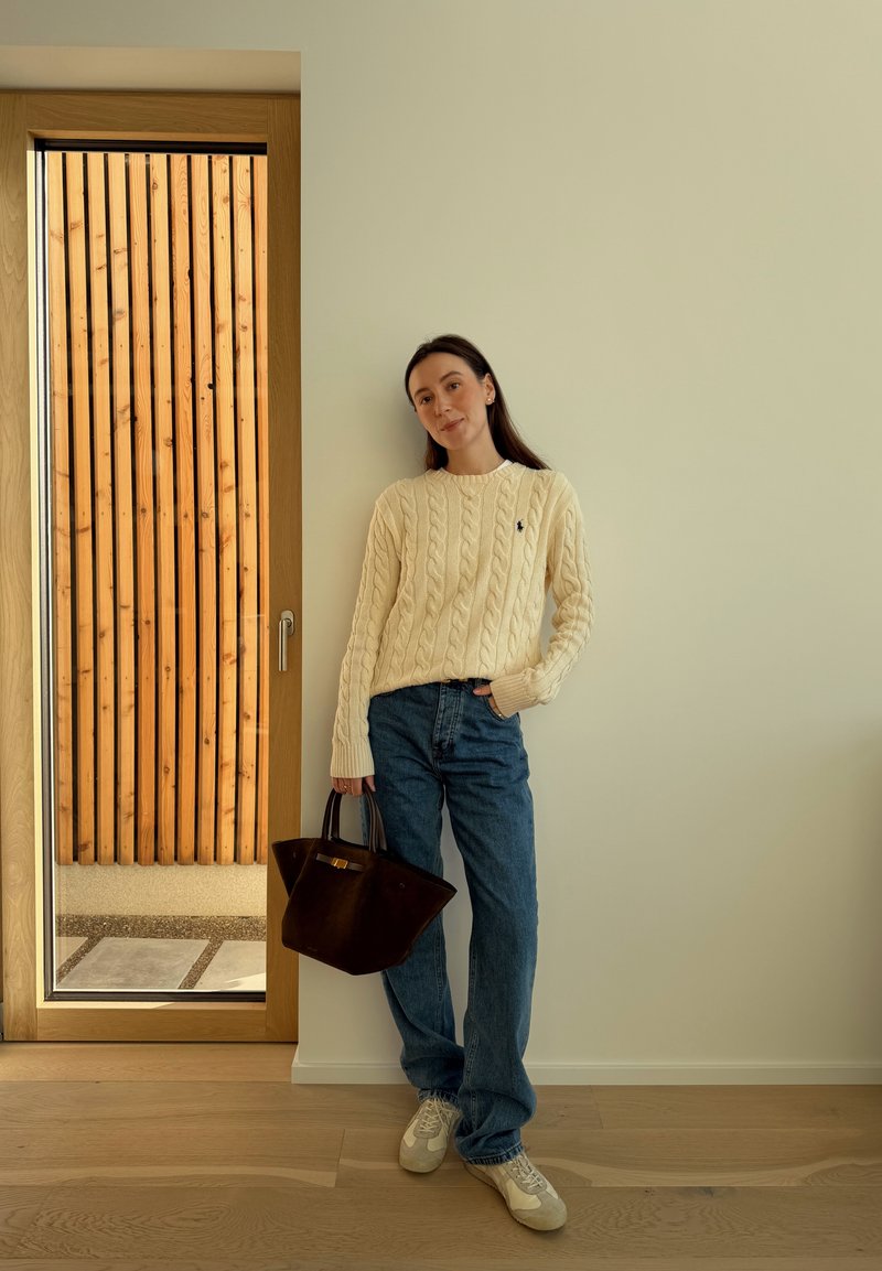 Femme appuyée contre un mur beige, portant un pull en maille torsadée crème, un jean bleu, des baskets blanches, tenant un grand sac à main marron foncé.