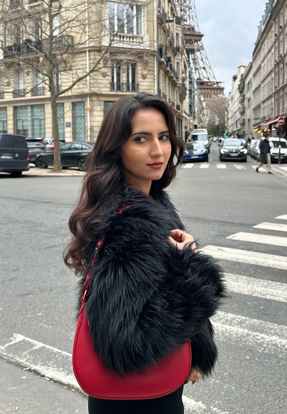 Rød læderhandtaske med en buet form og en aftagelig rem, båret med en sort faux fur-frakke, mod en urban baggrund.