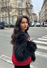 Rød læderhandtaske med en buet form og en aftagelig rem, båret med en sort faux fur-frakke, mod en urban baggrund.