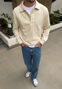 Pullover de cor creme com um decote com gola e três botões, combinado com jeans azuis de perna reta e ténis brancos.