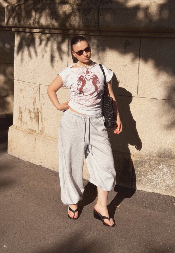 Frau mit Sonnenbrille, weißem bedrucktem T-Shirt, lockerer grauer Hose und schwarzen Sandalen steht an einer sonnenbeschienenen Steinmauer und trägt eine schwarze geflochtene Tasche.