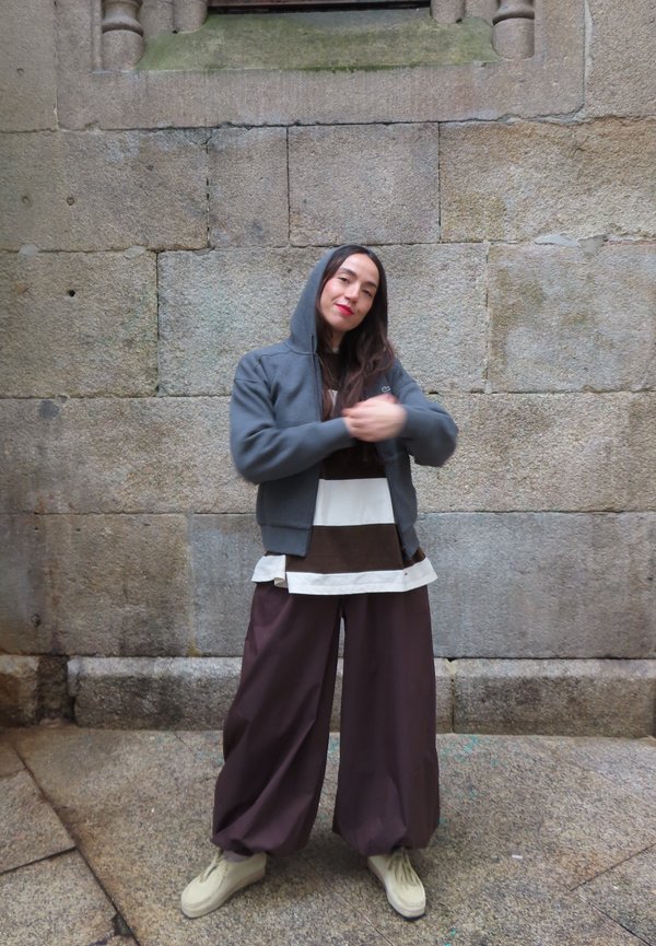 Personne portant un sweat à capuche gris, une chemise rayée, un pantalon large marron et des chaussures beige, debout contre un mur en pierre en train de relever sa manche.