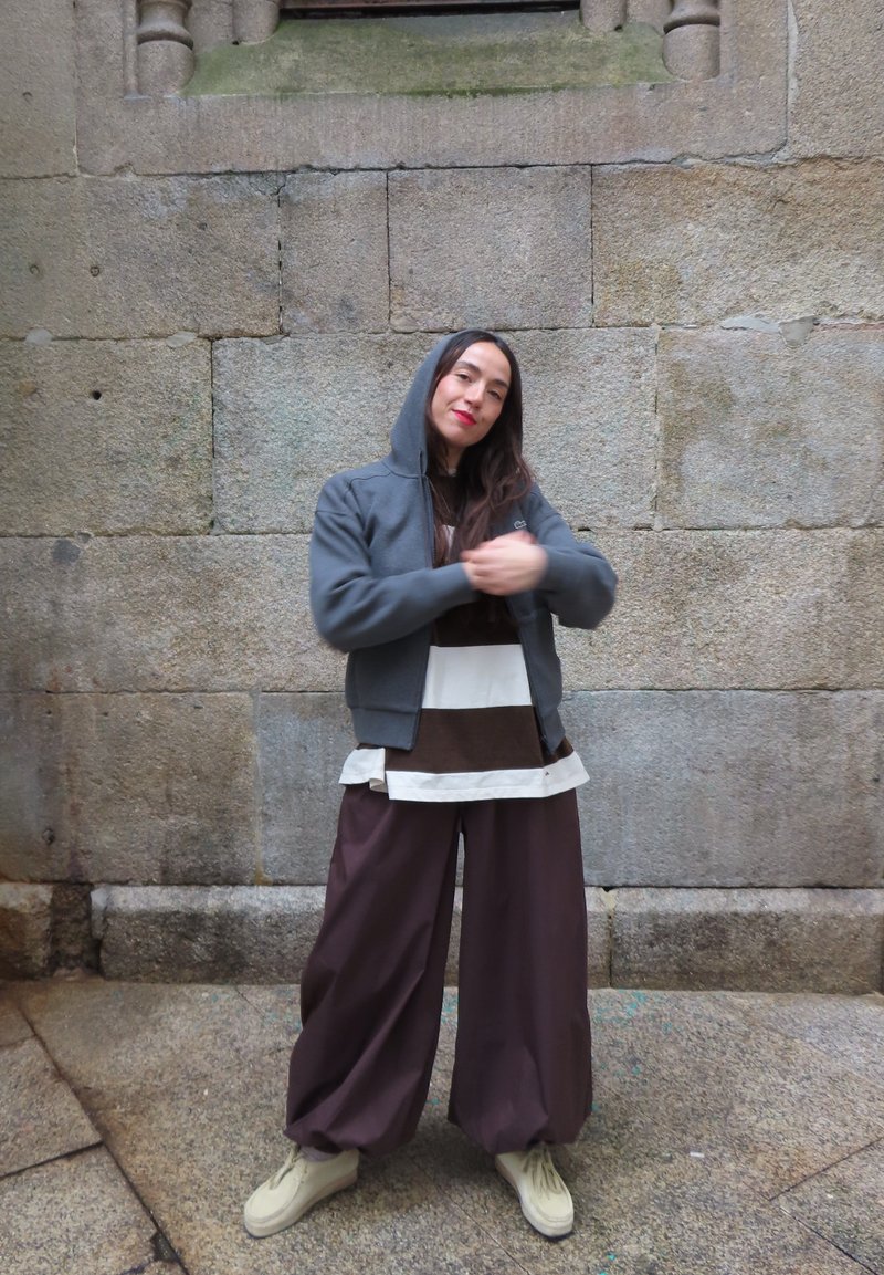 Person mit grauem Kapuzenpulli, gestreiftem Hemd, weiten braunen Hosen und beigen Schuhen steht an einer Steinmauer und zieht an seinem Ärmel.