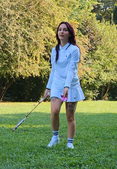 Camisa polo blanca con cuello, falda blanca con pliegues, calcetas de rayas blancas y negras, sosteniendo una raqueta de bádminton y un objeto rosa en un parque.