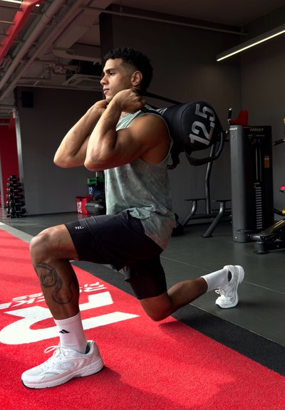 Bolsa de arena negra con un peso de 12,5 kg sobre los hombros de una persona que lleva una camiseta sin mangas gris claro y pantalones cortos negros, sobre una colchoneta roja de gimnasio.