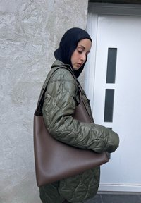 Καφέ δερμάτινη τσάντα tote με λεία επιφάνεια, φαρδιά βάση και μαλακές, στρογγυλεμένες άκρες, που κρατάει ένα άτομο ντυμένο με μια ελαιοπράσινη τζάκετ με επένδυση.