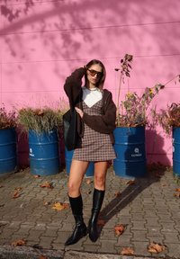 Smeđi oversized džemper, bijela majica, karirana haljina, crne čizme do koljena. U pozadini plavi bačve i ružičasti zid s biljkama.