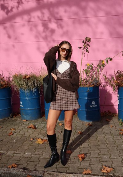 Suéter marrón holgado, top blanco, vestido de cuadros, botas negras hasta la rodilla. Fondo con barriles azules y una pared rosa con plantas.