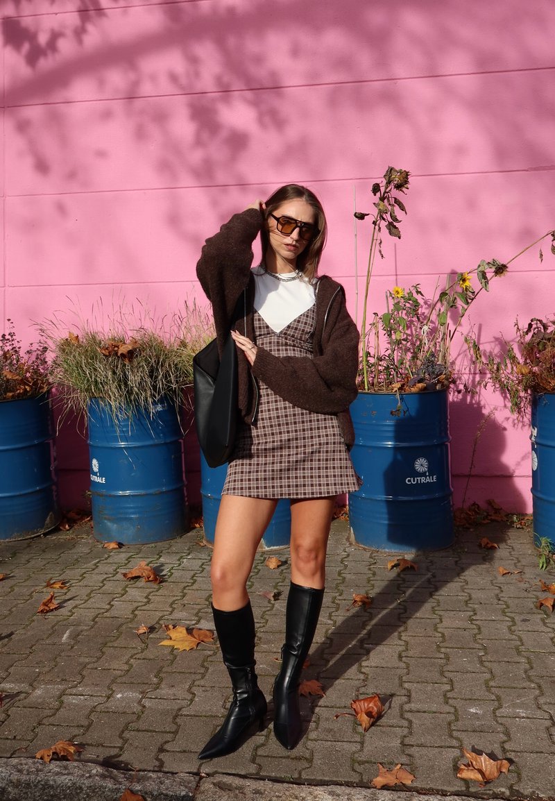 Suéter castanho oversized, top branco, vestido de padrão xadrez, botas pretas até ao joelho. Fundo com tambores azuis e uma parede rosa com plantas.