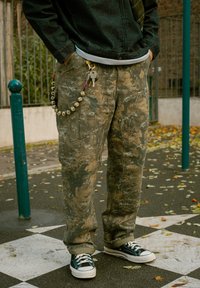 Pantaloni cargo camuflați, cu croială largă, accesorizați cu un breloc format din mărgele și chei. Purtate cu adidași negri și albi.