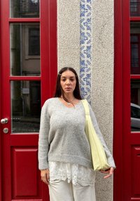 Νεαρή γυναίκα με μακριά σκούρα μαλλιά στέκεται μπροστά από κόκκινες πόρτες, φοράει γκρι πουλόβερ, λευκή δαντελένια μπλούζα και κρατά κίτρινη τσάντα ώμου.