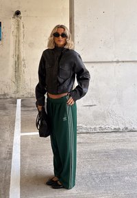 Kvinne med blondt hår iført svart kort skinnjakke, mørke solbriller, grønne Adidas treningsbukser, holder en svart håndveske i et parkeringshus.