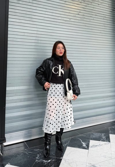 Mujer con chaqueta de cuero negra, suéter negro con logo blanco, falda blanca con lunares, botas negras, sosteniendo un bolso blanco, de pie junto a una persiana metálica.