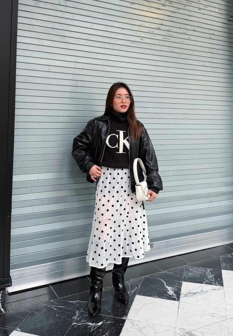 Frau trägt schwarze Lederjacke, schwarzen Pullover mit weißem Logo, weißen Rock mit Punkten, schwarze Stiefel, hält eine weiße Handtasche und steht vor einem Metallrollladen.