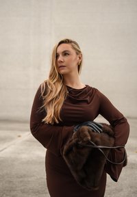 Brun kjole med draperinger og klokkeærmer, båret med sorte handsker og en brun pels-håndtaske. Langt, bølget hår og minimal smykker fremhæver looket.