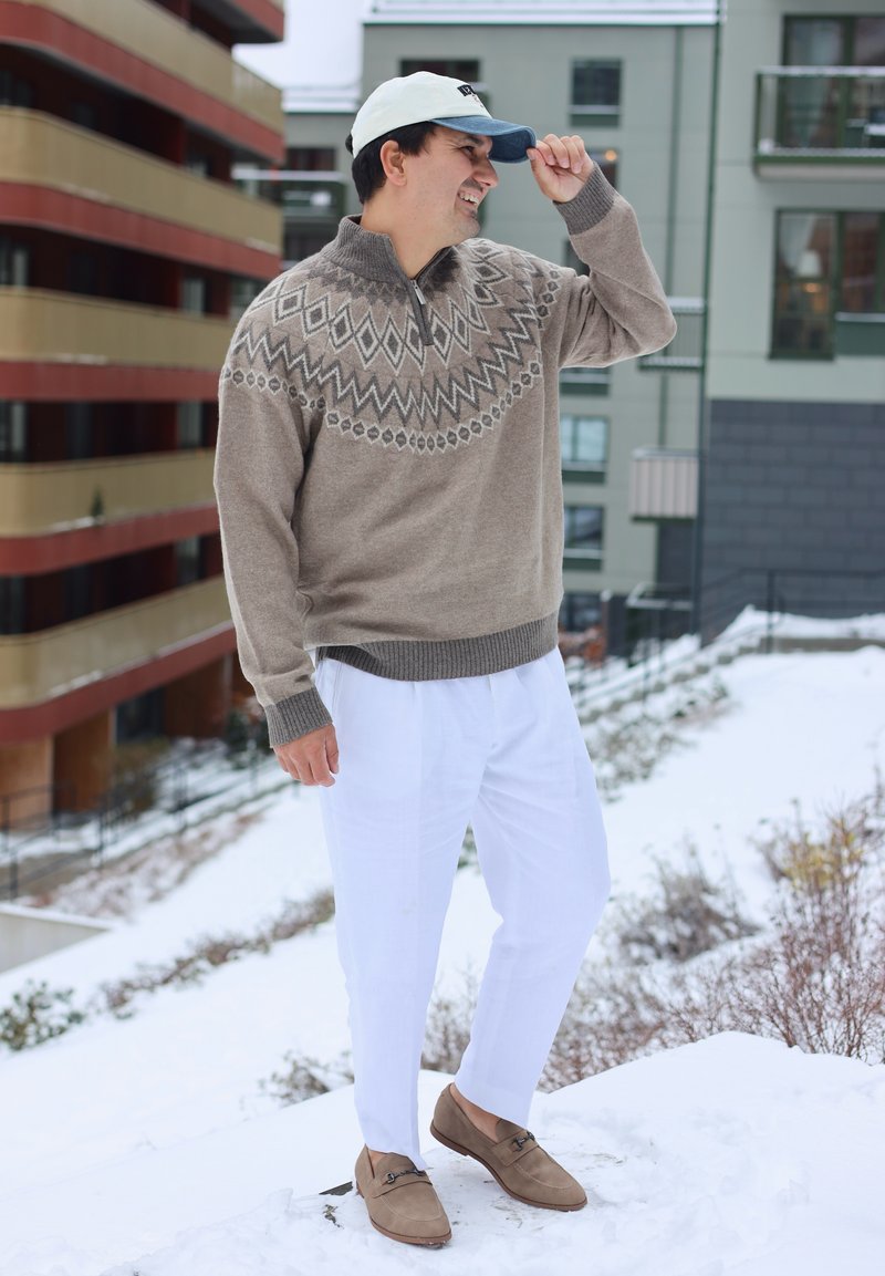 Homme portant un pull beige à motifs, un pantalon blanc et une casquette, debout à l'extérieur sur la neige près d'immeubles d'habitation.