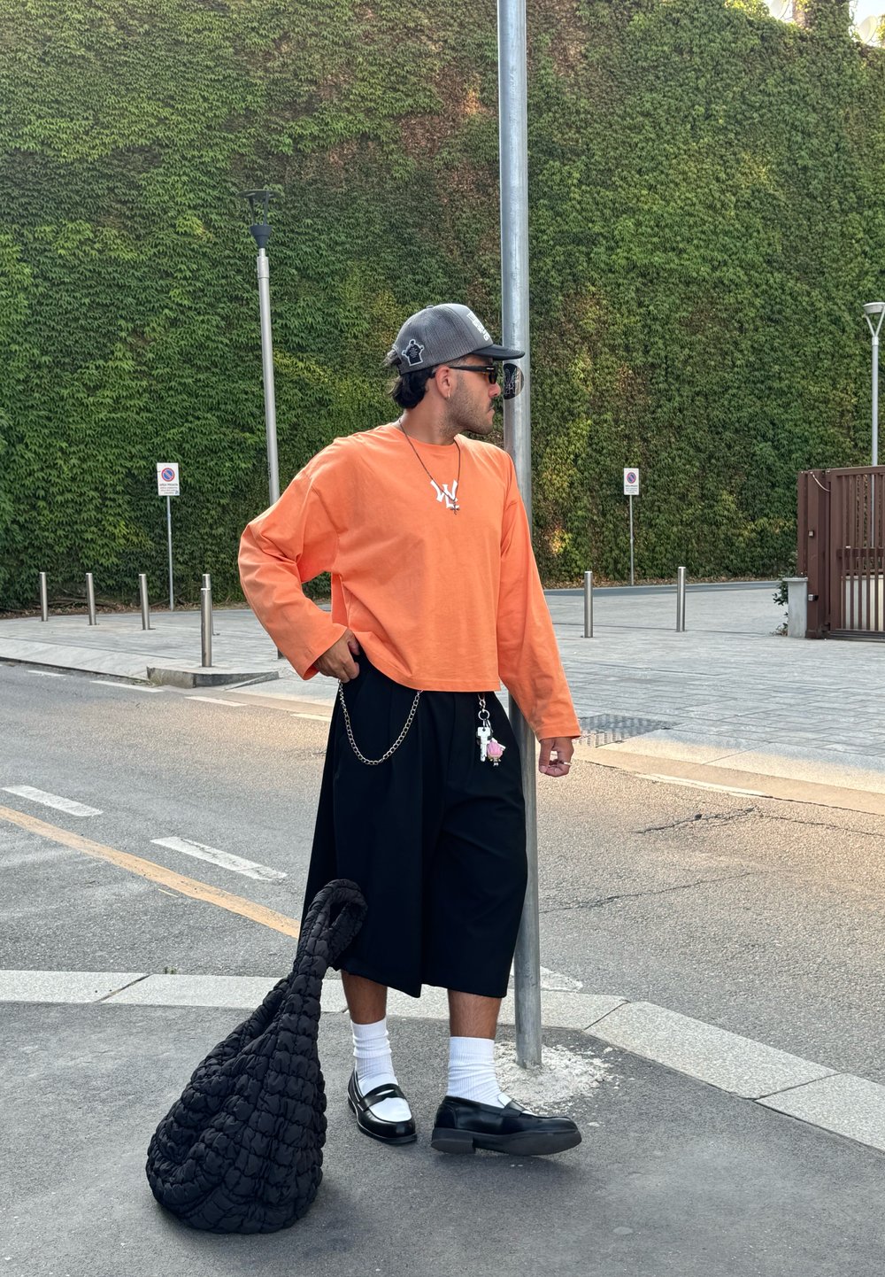 Oranje lange mouwen shirt, zwarte wijde shorts met kettingdetail, witte sokken en zwarte schoenen. Zwarte gestructureerde tas gepositioneerd bij de voeten.