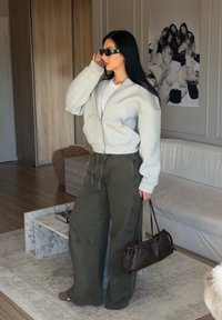 Corta felpa grigia chiara con zip, t-shirt bianca sotto, pantaloni verdi larghi e una borsa a tracolla in pelle marrone, abbinati a occhiali da sole neri.