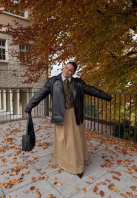 Svart skinn bikerjakke, grønn genser, mønstret slips, beige maksi-skjørt, svart håndveske, står på steinfliser med høstblader i bakgrunnen.