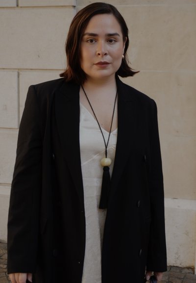 Abrigo largo negro sobre un vestido de satén crema, con un collar de borlas negras y una cuenta redonda. Textura suave, diseño estructurado.