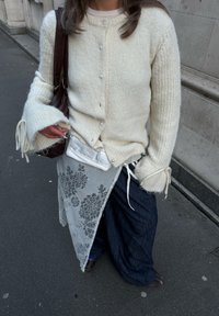 Cardigan en maille blanc avec de gros boutons, associé à une surcouche transparente brodée de fleurs et un pantalon ample bleu foncé. Sac à main marron inclus.