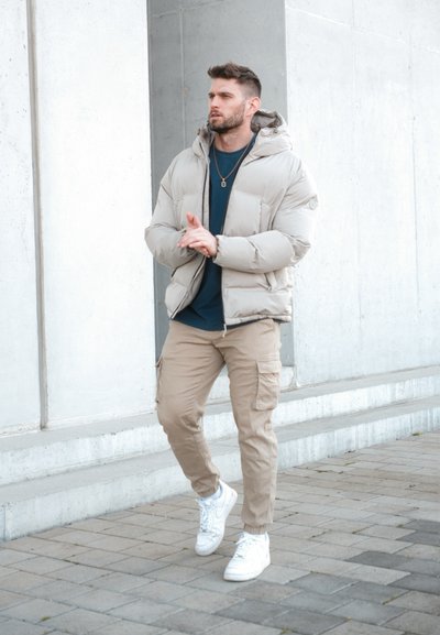 Chaqueta acolchada color beige con capucha, combinada con una camiseta azul marino y pantalones cargo beige. Zapatillas blancas, diseño minimalista y corte relajado.