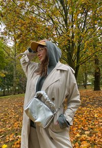 Bež trench coat z zategnjenim pasom, sivo kapuco pod njim in srebrna kovinska torbica. Ozadje prikazuje jesensko listje z oranžnimi listi.