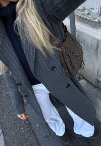 Hall triibuline mantel mustade nuppudega, tumesinine kõrge kaelusega kampsun, valged laiade jalgadega püksid ja pruunist mustriga õlakott, kantud pruuni jalatsitega.