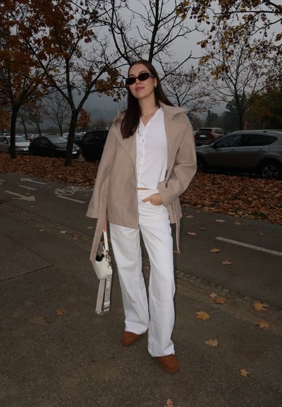 Abrigo beige de lana, camisa blanca abotonada, pantalones blancos de pierna ancha, zapatos marrones y un bolso pequeño blanco con correa. Escenario al aire libre con hojas otoñales.