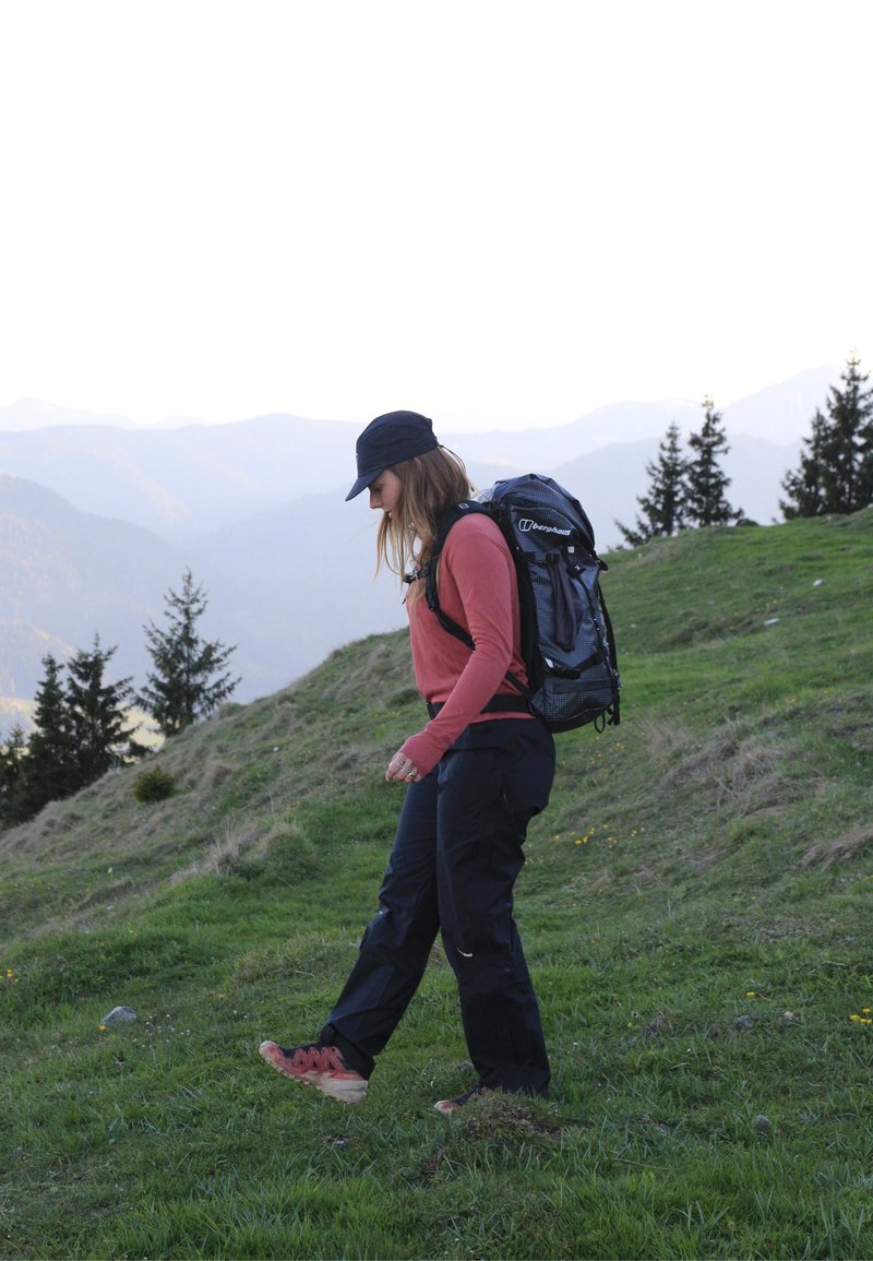 Femme qui fait de la randonnée sur un versant de montagne herbeux, portant un sac à dos, une casquette et des vêtements d'extérieur, avec des arbres et des montagnes lointaines en arrière-plan.