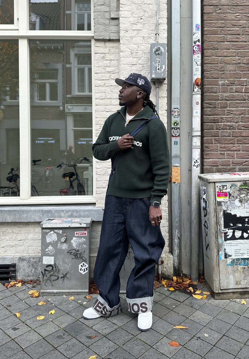 Muž v tmavom svetri a čiapke, široké manžetové džínsy s textom, biele tenisky, stojaci na chodníku pokrytom lístím pri tehlovej stene a rozvodných skriniach.
