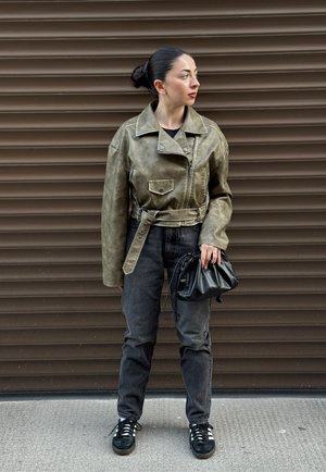 Chaqueta de cuero verde oliva con cintura ceñida y gran solapa, combinada con jeans negros y un pequeño bolso negro. Unas zapatillas negras completan el look.