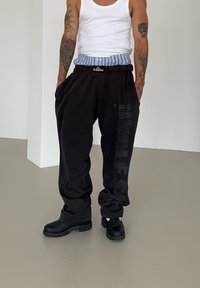 Calças de treino pretas com tornozelos estreitados e texto impresso na perna esquerda, combinadas com tamancos pretos e uma t-shirt branca.