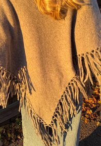Poncho marron à franges en tissu doux et texturé, de forme triangulaire, avec de longues pampilles sur les bords.