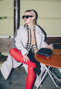 Béžový trenčkot s černými pruhy, červené punčocháče, černé boty a šála s Burberry vzorem. Černá kabelka s řetízkovým detailem na kulatém stole.