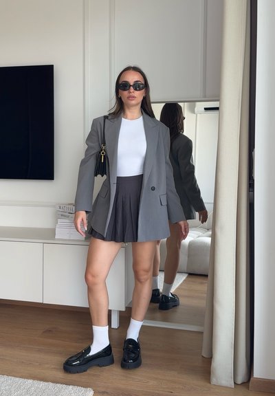 Mujer con gafas de sol, blazer gris, top blanco, falda oscura plisada, calcetines blancos, mocasines negros, de pie en una habitación con espejo y televisión.