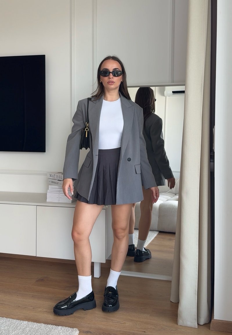 Femme portant des lunettes de soleil, blazer gris, haut blanc, jupe plissée foncée, chaussettes blanches, mocassins noirs, debout dans une pièce avec un miroir et une télévision.