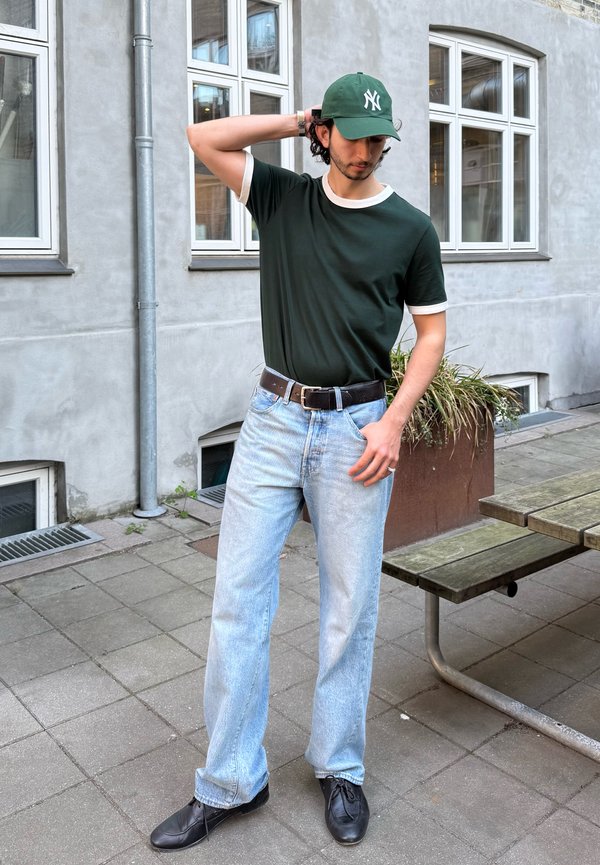 Bărbat purtând șapcă verde și ochelari cu ramă neagră, tricou verde închis, blugi bleu deschis și pantofi negri, stând în aer liber aproape de o clădire și o bancă.