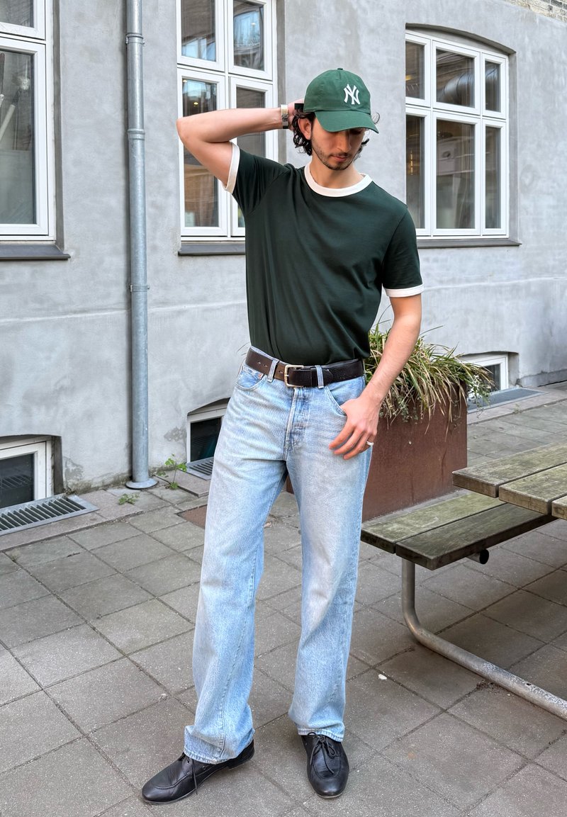 Homme portant une casquette verte et des lunettes à monture noire, un t-shirt vert foncé, un jean bleu clair et des chaussures noires, debout à l'extérieur près d'un bâtiment et d'un banc.