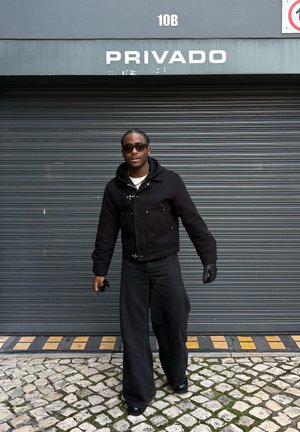 Hombre con gafas de sol negras, chaqueta, guantes y pantalones anchos caminando sobre adoquines delante de una puerta enrollable gris con el texto "PRIVADO" arriba.