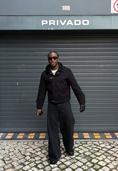 Hombre con gafas de sol negras, chaqueta, guantes y pantalones anchos caminando sobre adoquines delante de una puerta enrollable gris con el texto "PRIVADO" arriba.